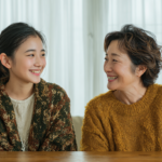 Two women sit at a wooden table indoors, smiling warmly at each other as they chat in cozy knitwear.