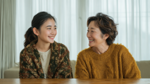 Two women sit at a wooden table indoors, smiling warmly at each other as they chat in cozy knitwear.