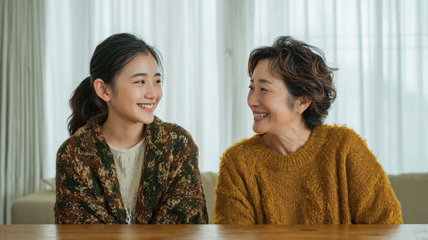 Two women sit at a wooden table indoors, smiling warmly at each other as they chat in cozy knitwear.