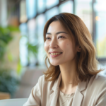 Smiling woman in a light blazer sits in a bright cafe with greenery in the background and large windows nearby.