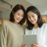 Two women smiles at a tablet, standing together in a bright indoor space like a cafe or shop.