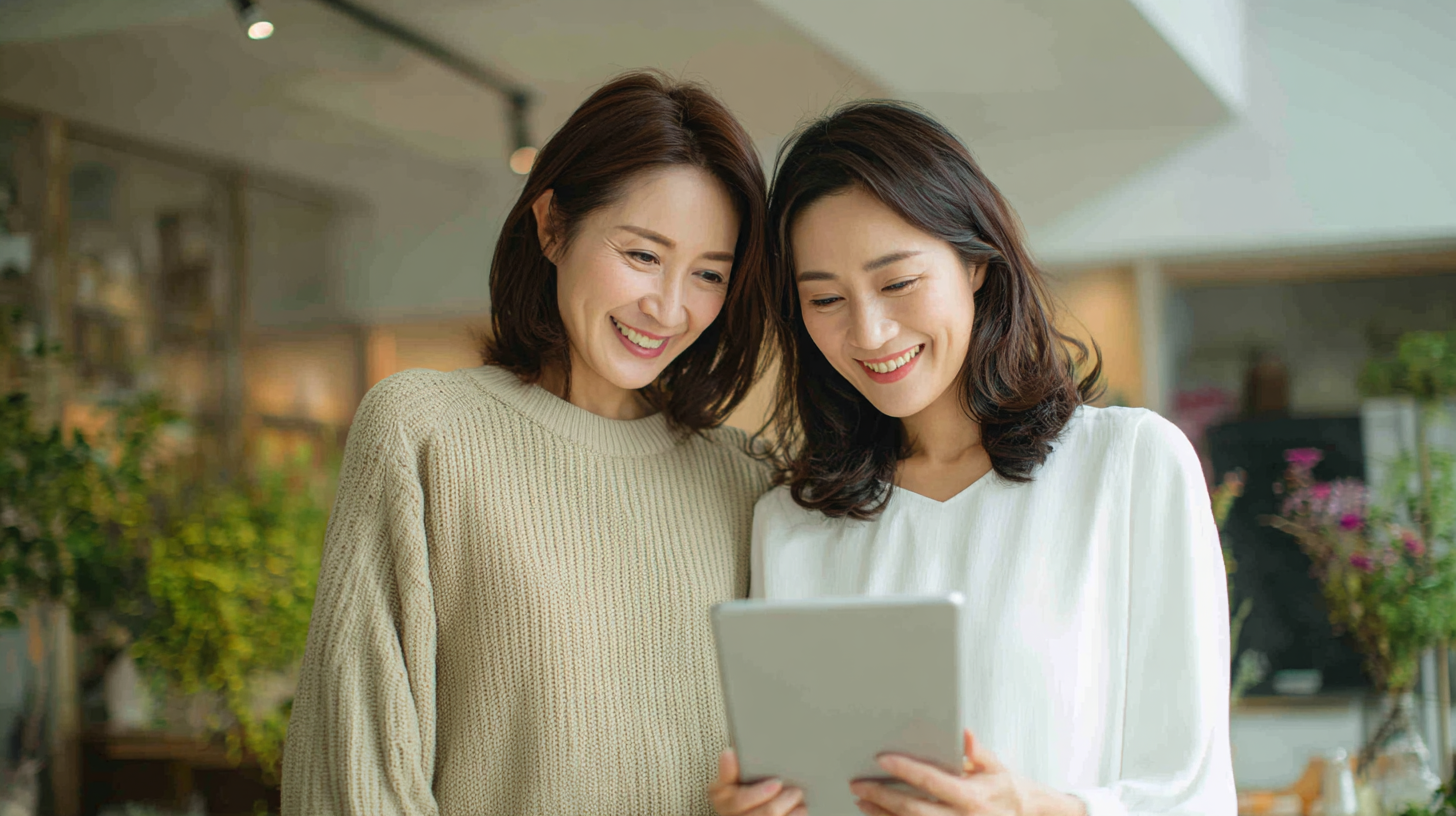 Two women smiles at a tablet, standing together in a bright indoor space like a cafe or shop.