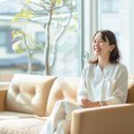 Smiling woman in a white uniform sitting on a beige sofa in a sunlit room with large windows and plants behind her.