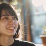 Portrait of a smiling young woman with short black hair, bathed in warm sunlight indoors, looking up slightly to the left.