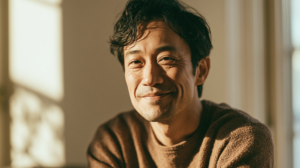 Close-up portrait of a smiling Asian man with dark hair in a brown knit sweater, bathed in warm indoor sunlight.