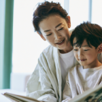 Adult and child reading a book together on a sofa by large windows, smiling.