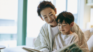 Adult and child reading a book together on a sofa by large windows, smiling.