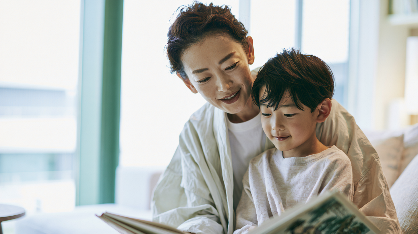 Adult and child reading a book together on a sofa by large windows, smiling.