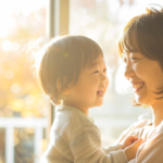 Mother holding a toddler, both smiling warmly at each other by a sunlit window.